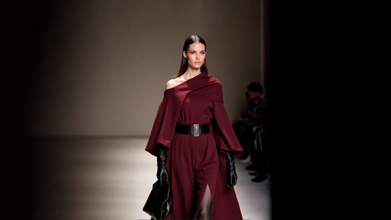 The model walks the runway wearing a cape-coat wit